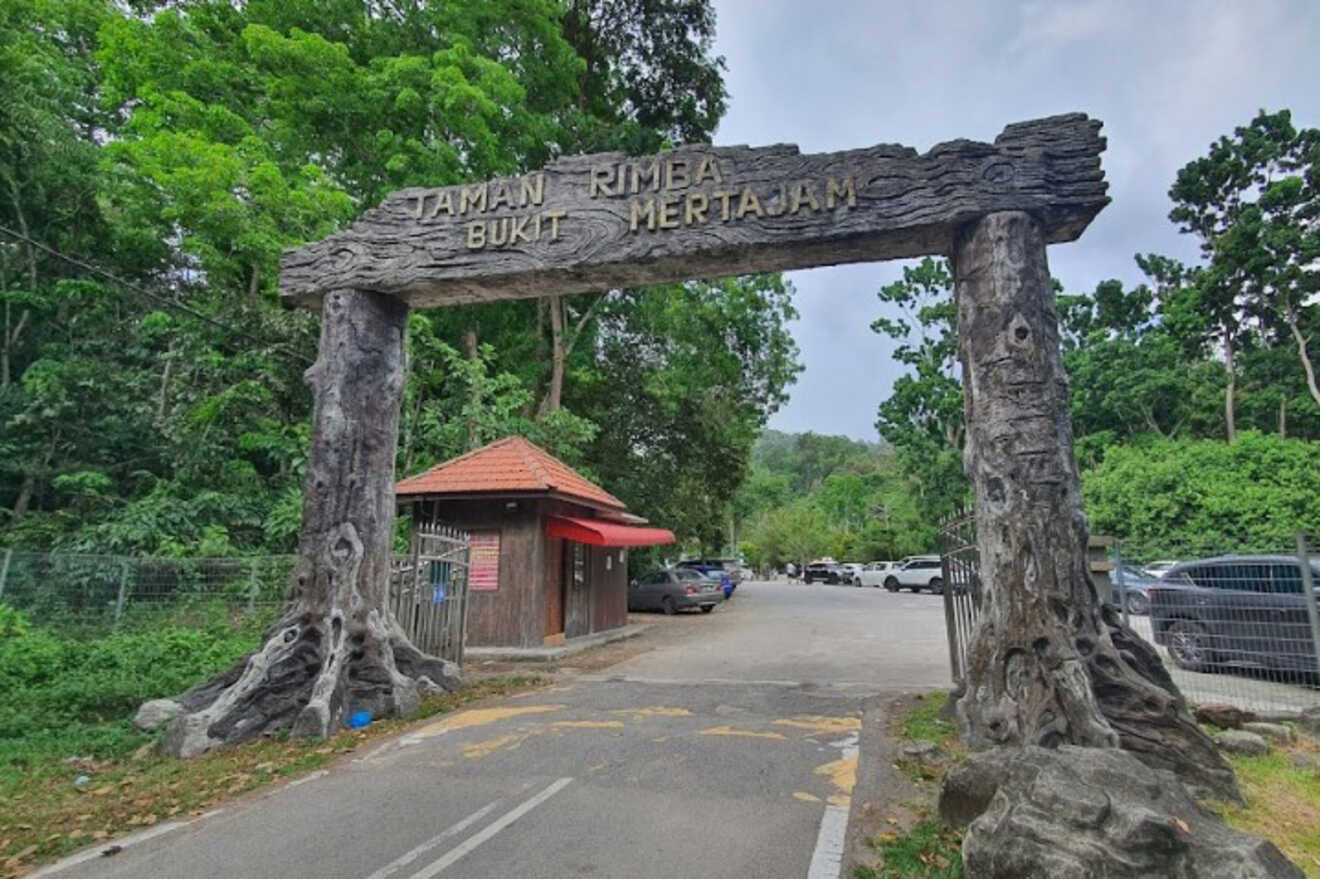 Entrance gate to Taman Rimba Bukit Mertajam, featuring a rustic wooden archway. A small building with a red roof is visible on the left, and cars are parked in the background. Lush greenery surrounds.