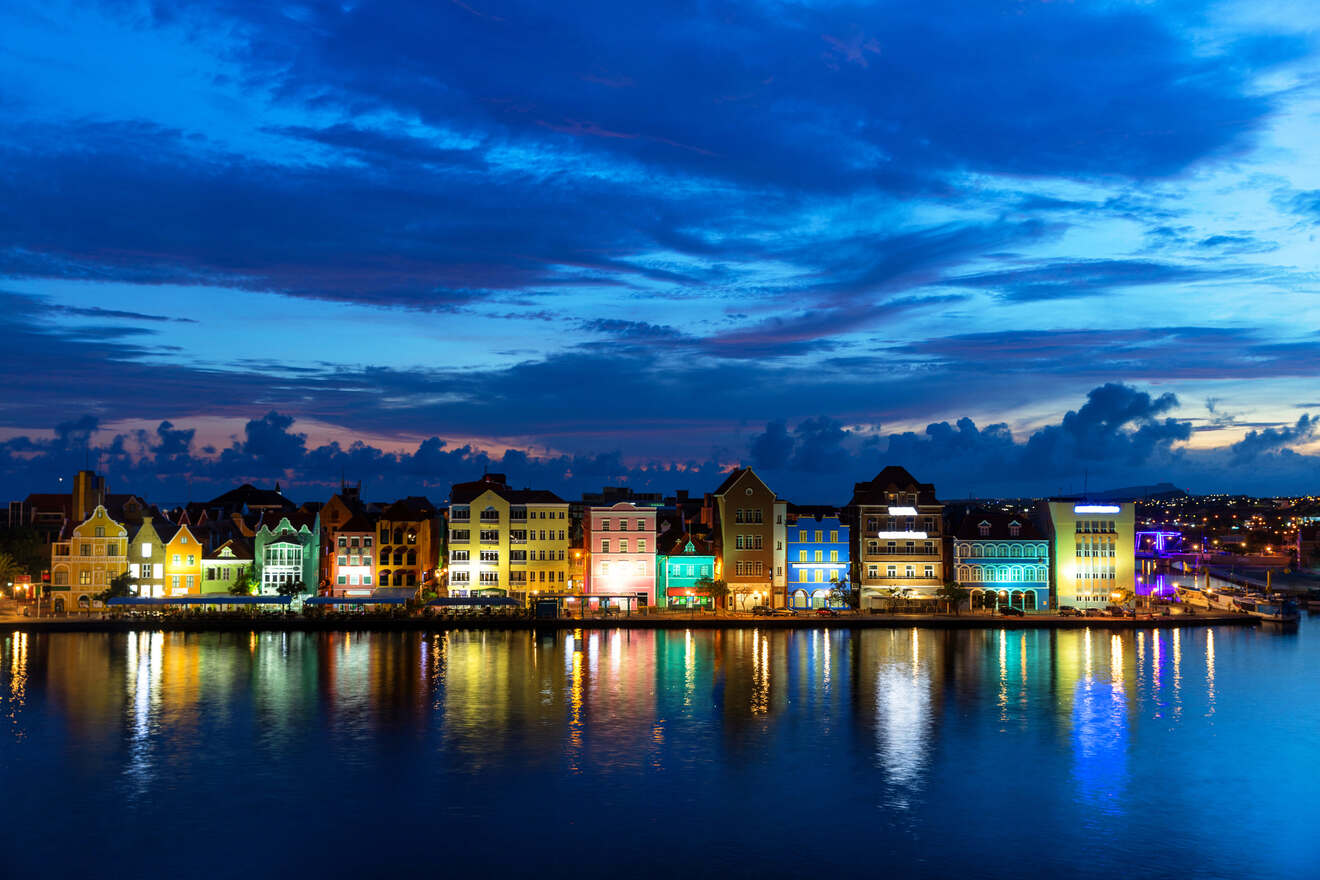 A row of colorful buildings along the waterfront is illuminated at dusk, with their reflections visible in the calm water below a partly cloudy, deep blue sky.