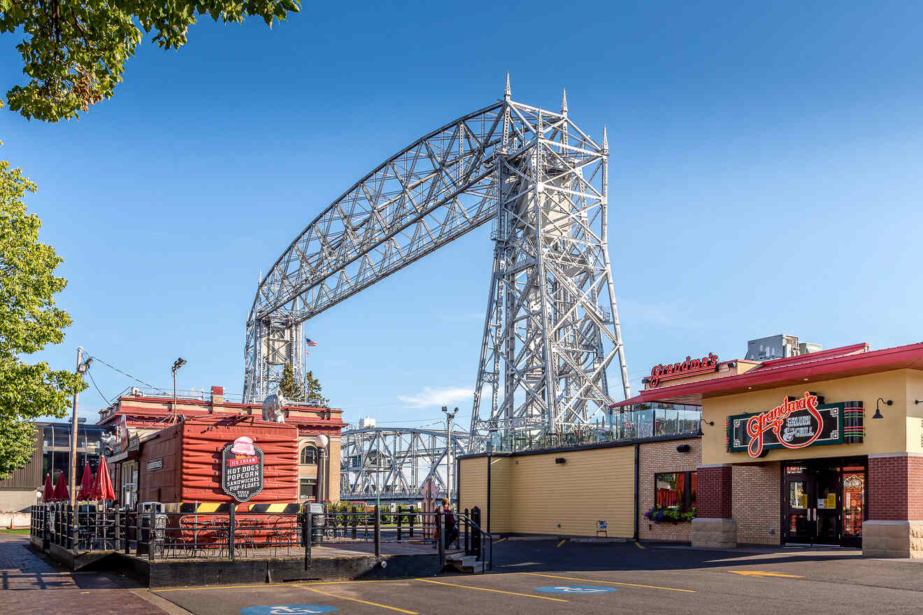 A large lift bridge over a clear blue sky is seen next to two restaurants with outdoor seating.