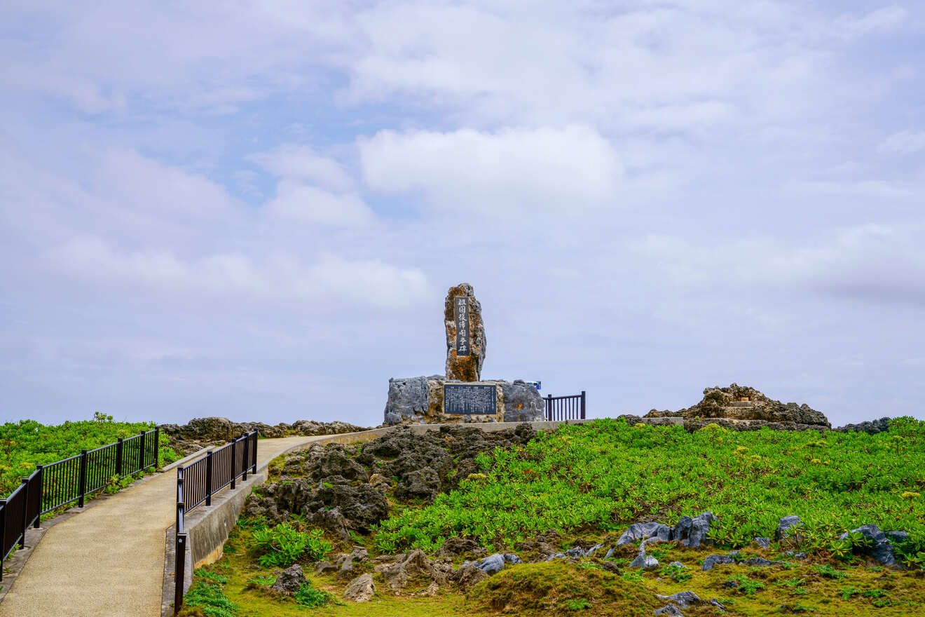 A stone monument stands at the end of a paved path surrounded by greenery and rocky terrain under a cloudy sky.