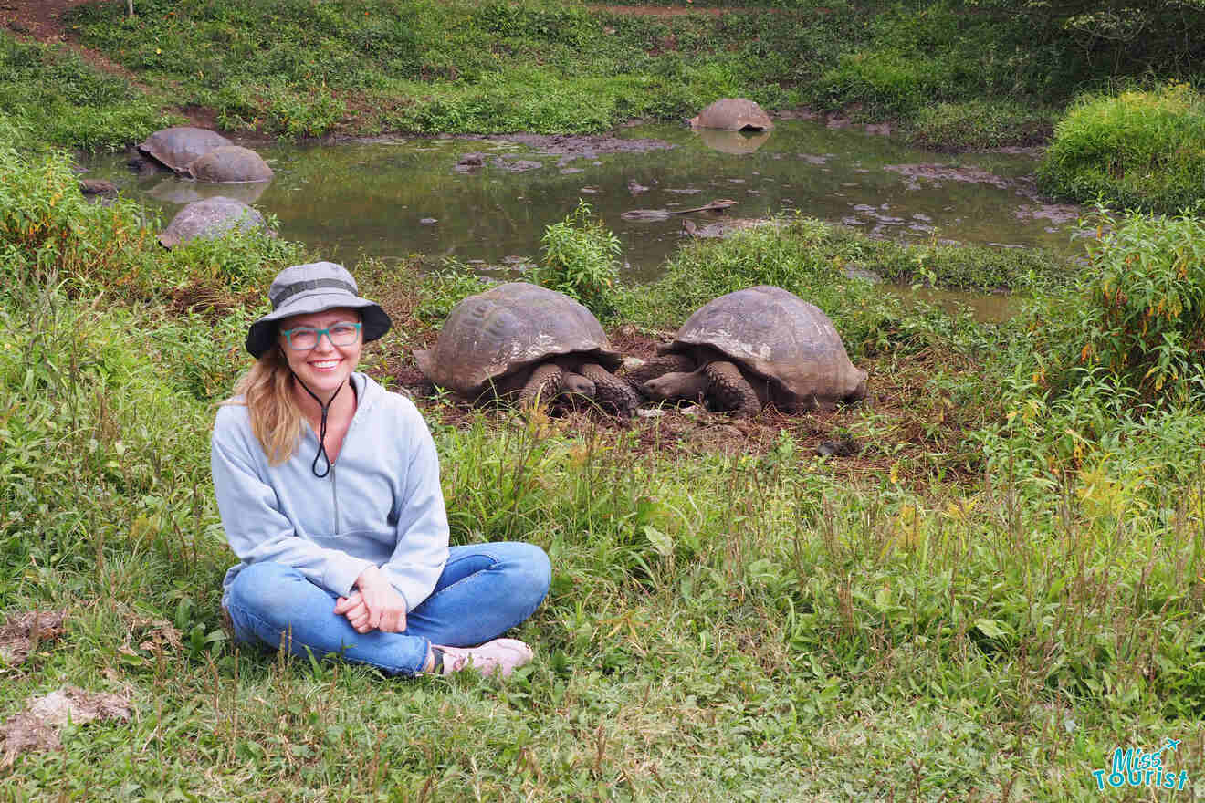 author of the post wearing a hat and glasses sits on the grass, smiling, with several large tortoises on a grassy area near a small pond in the background.