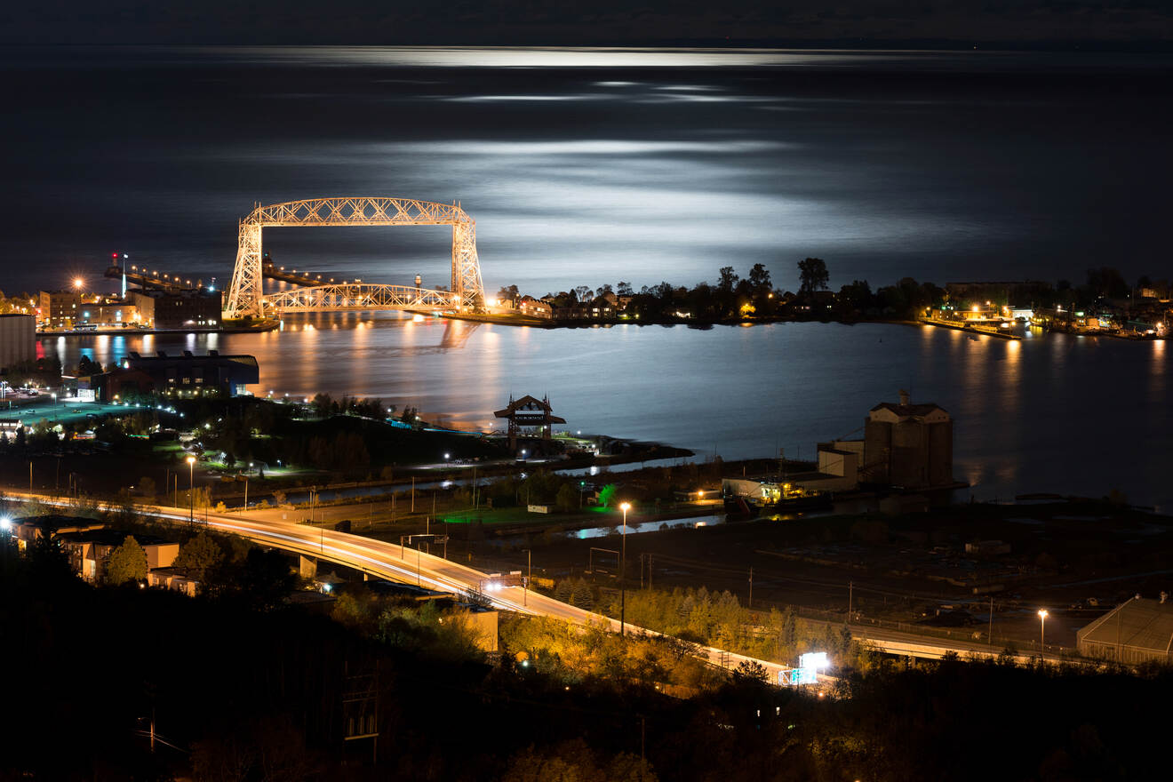 Night view of a lit-up bridge over a calm body of water, with a cityscape in the background and light reflecting on the water's surface.