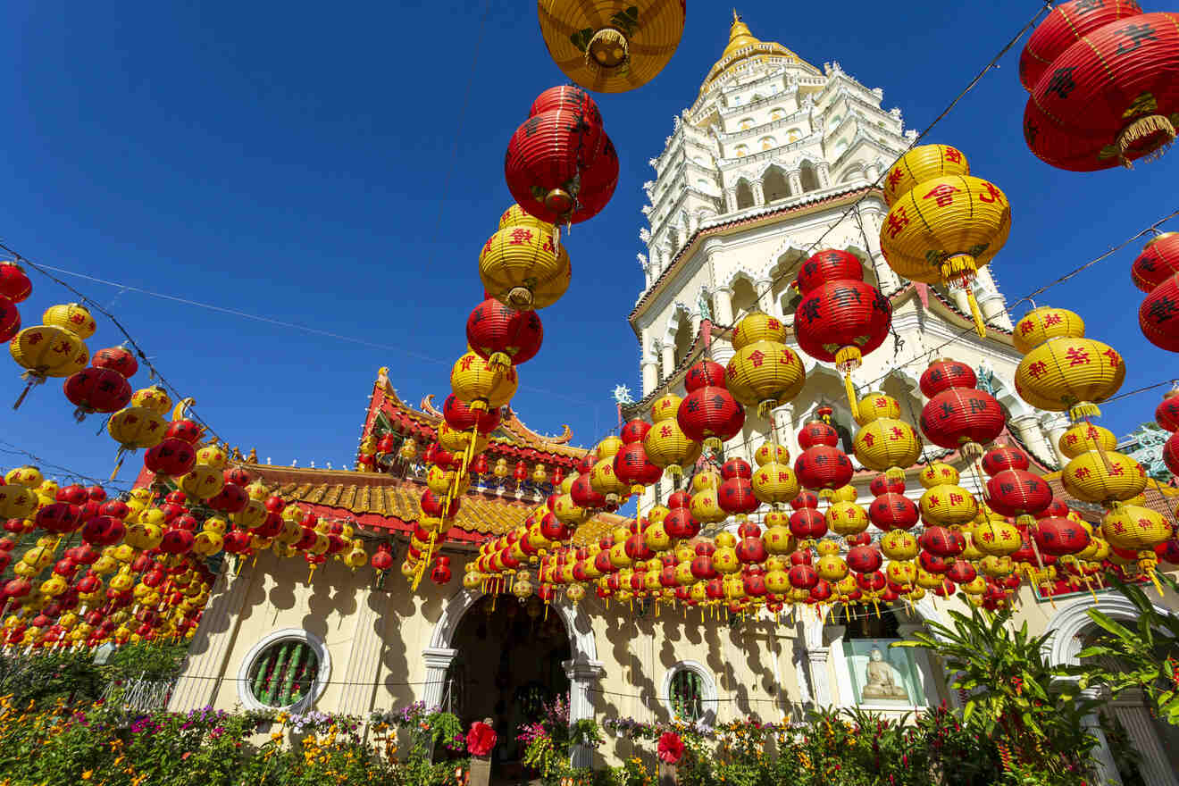 A temple with a tall white pagoda and an array of red and yellow lanterns hanging above, set against a clear blue sky.