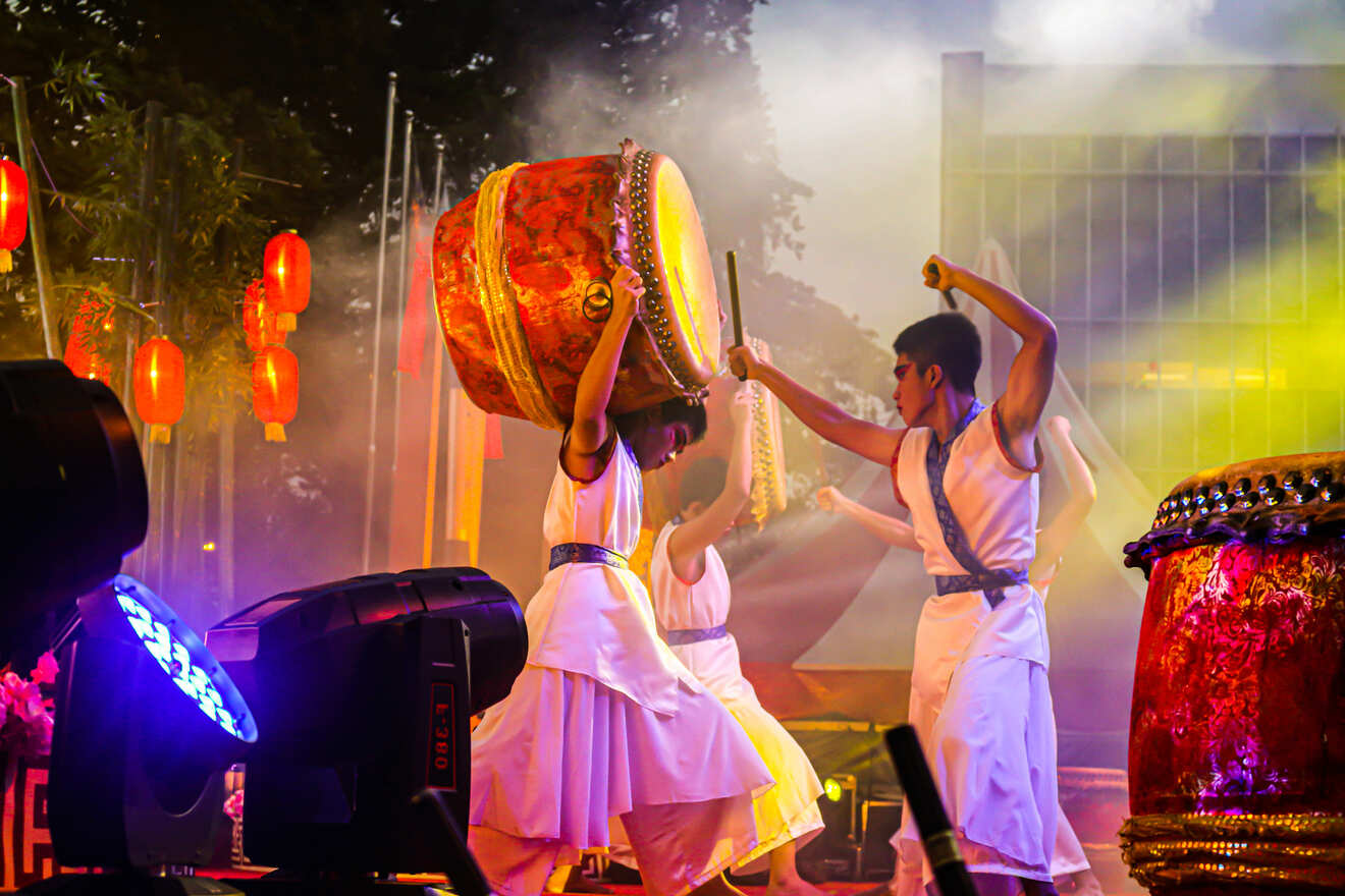 Performers dressed in white play large traditional drums on a stage with vibrant lighting and red lanterns in the background.