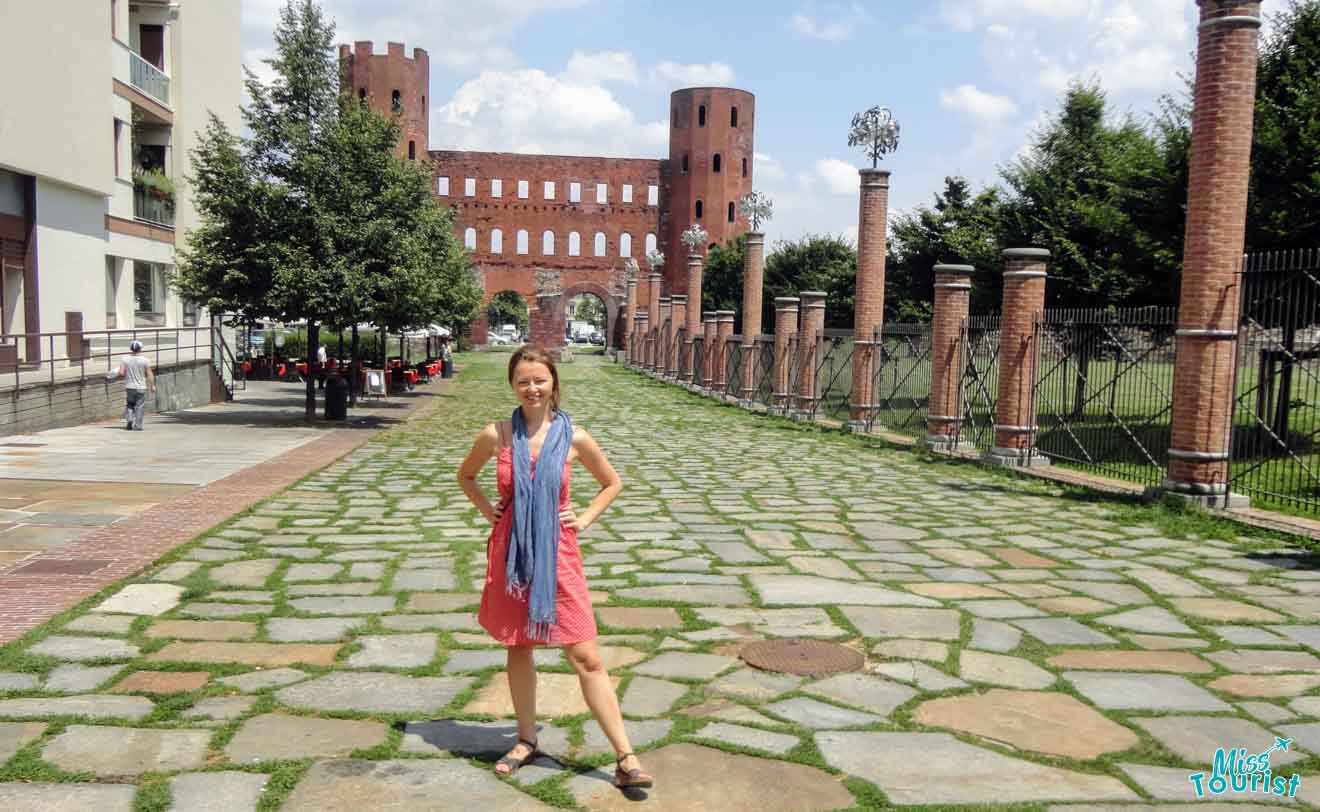 A tourist posing in front of the ancient Palatine Towers in Turin, reflecting the city's Roman history on a sunny day with clear skies.
