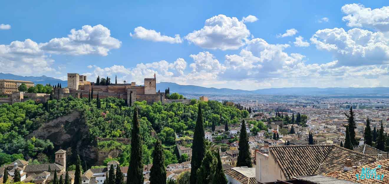 Breathtaking panoramic view of the Alhambra overlooking the cityscape of Granada, with lush greenery in the foreground and a backdrop of fluffy clouds against a blue sky