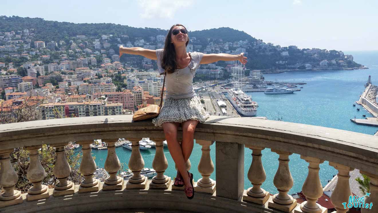 Yulia, the writer of the article with outstretched arms sitting on a balustrade with a panoramic view of Nice's coastline and port area in the background, under a clear blue sky.