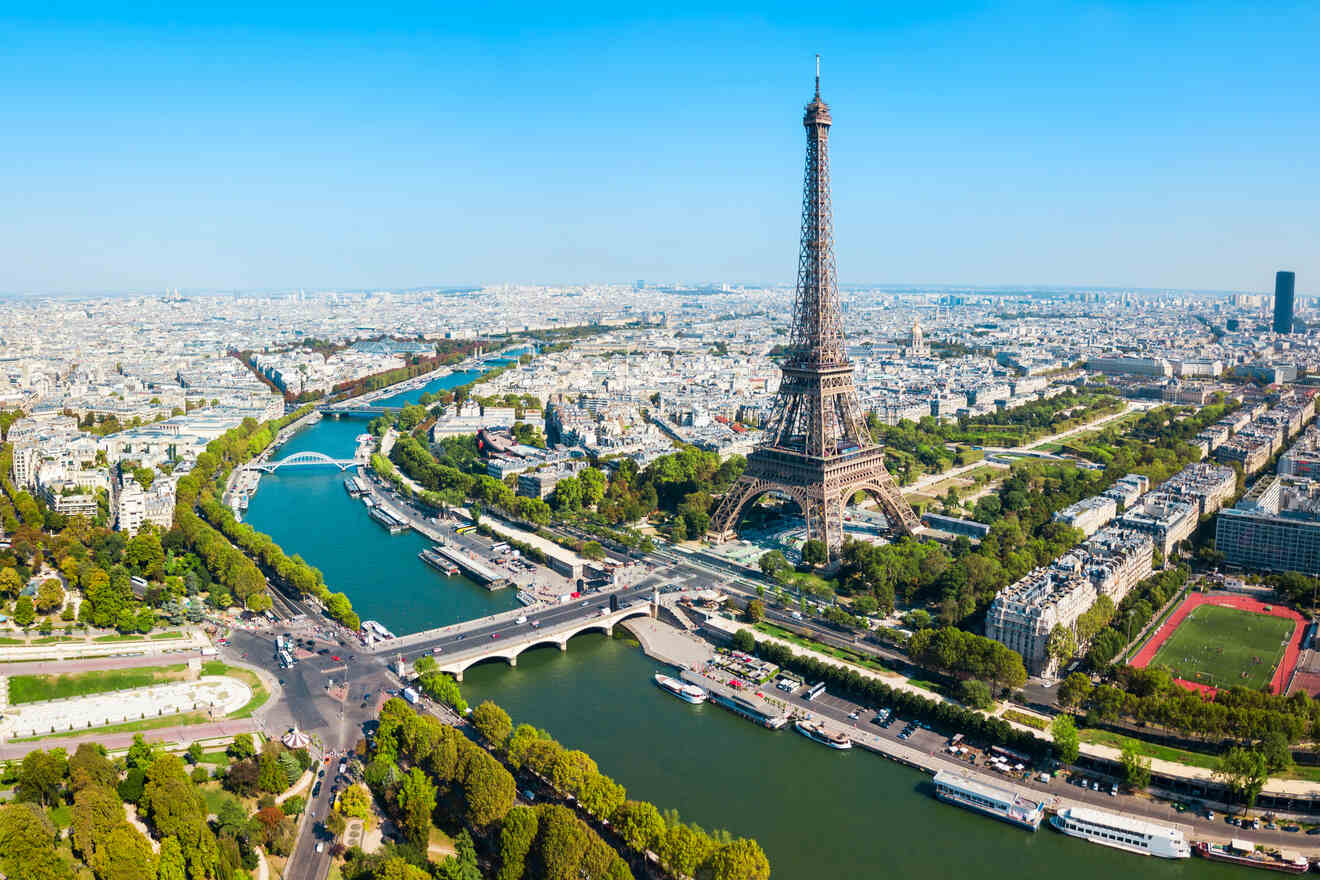 Aerial view of the Eiffel Tower in Paris, surrounded by city buildings, the Seine River with bridges, and green park areas under a clear blue sky.