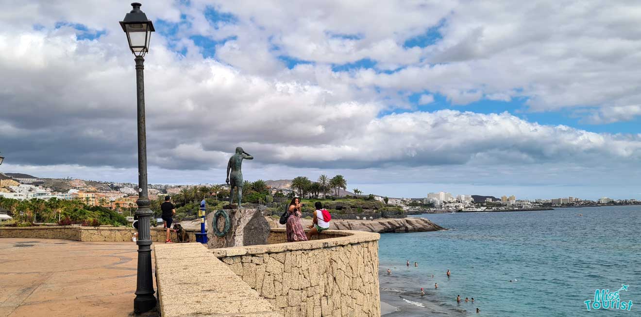 Picturesque seaside promenade with a bronze sculpture, tourists enjoying the view, and a panoramic view of the coastal town and cloudy sky