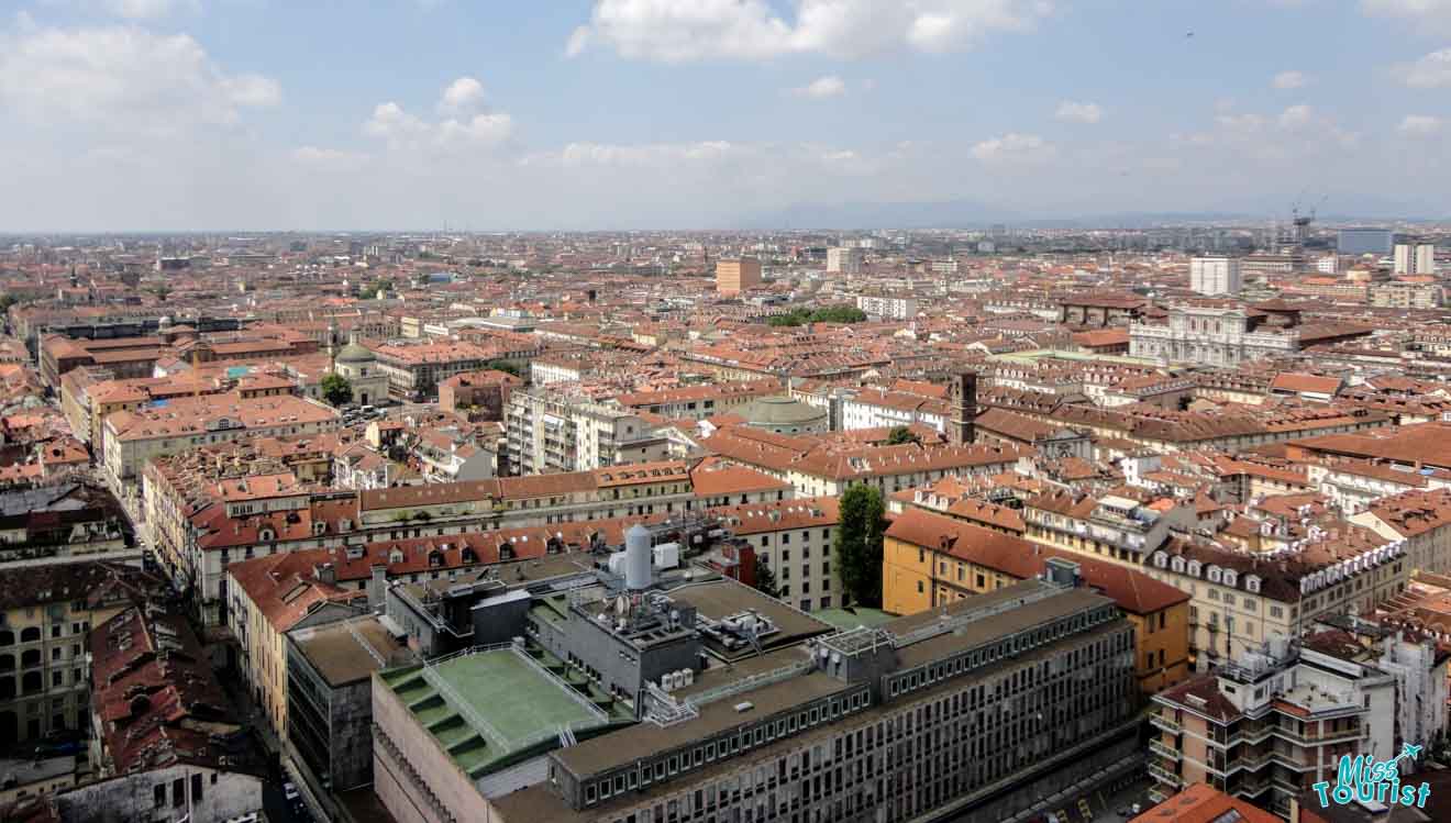 A panoramic view of Turin, Italy, displaying the dense urban landscape with historical buildings and modern structures under a cloudy sky.
