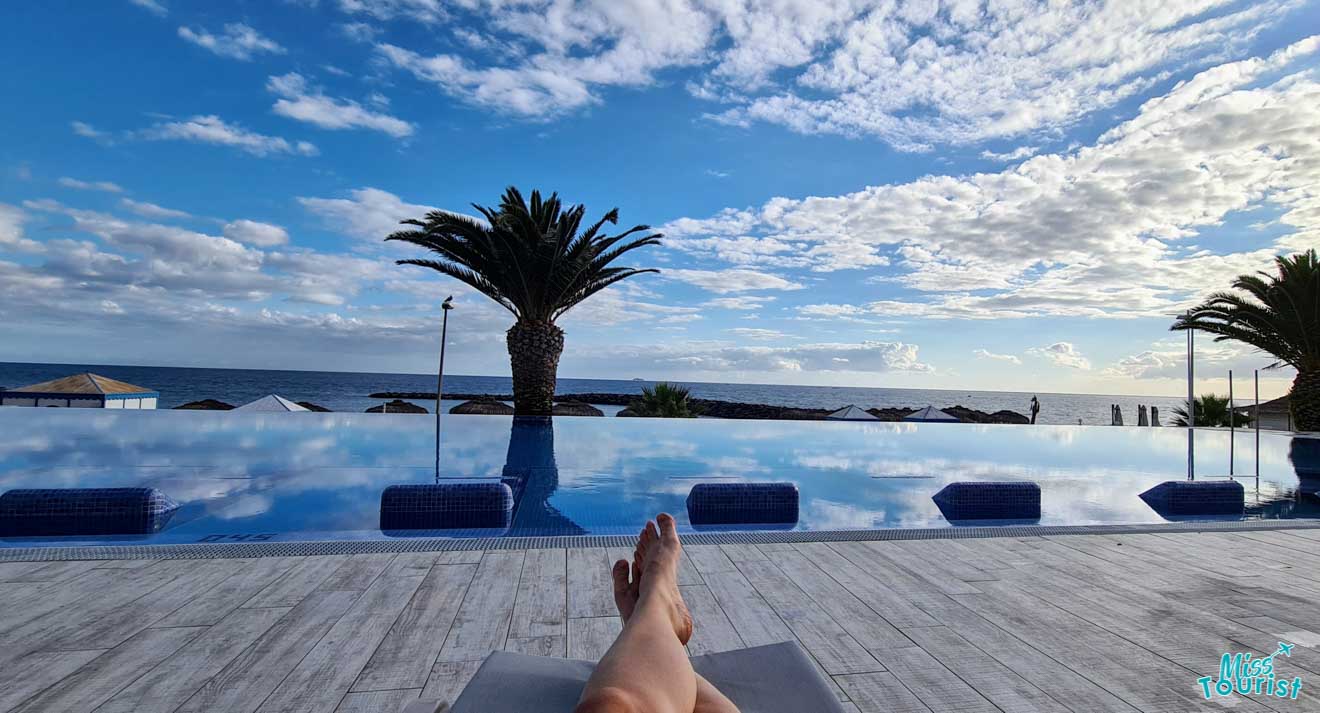 A first-person view of the writer of the post lounging by an infinity pool, overlooking the sea with a palm tree silhouette against a serene sky, embodying a moment of peaceful relaxation.