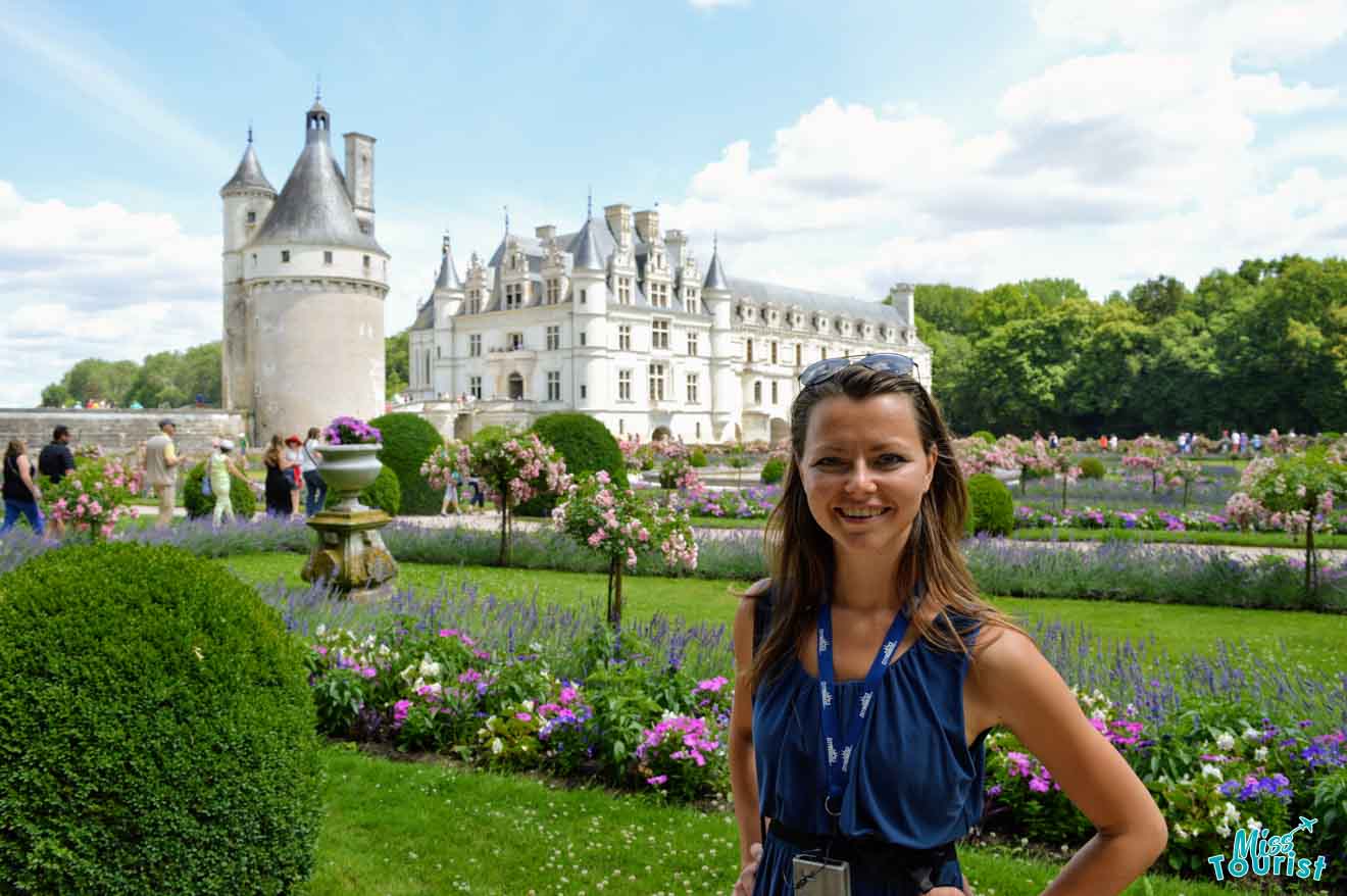 author of the post stands smiling in front of a lavish garden with a historic castle in the background. Other visitors are visible in the garden.