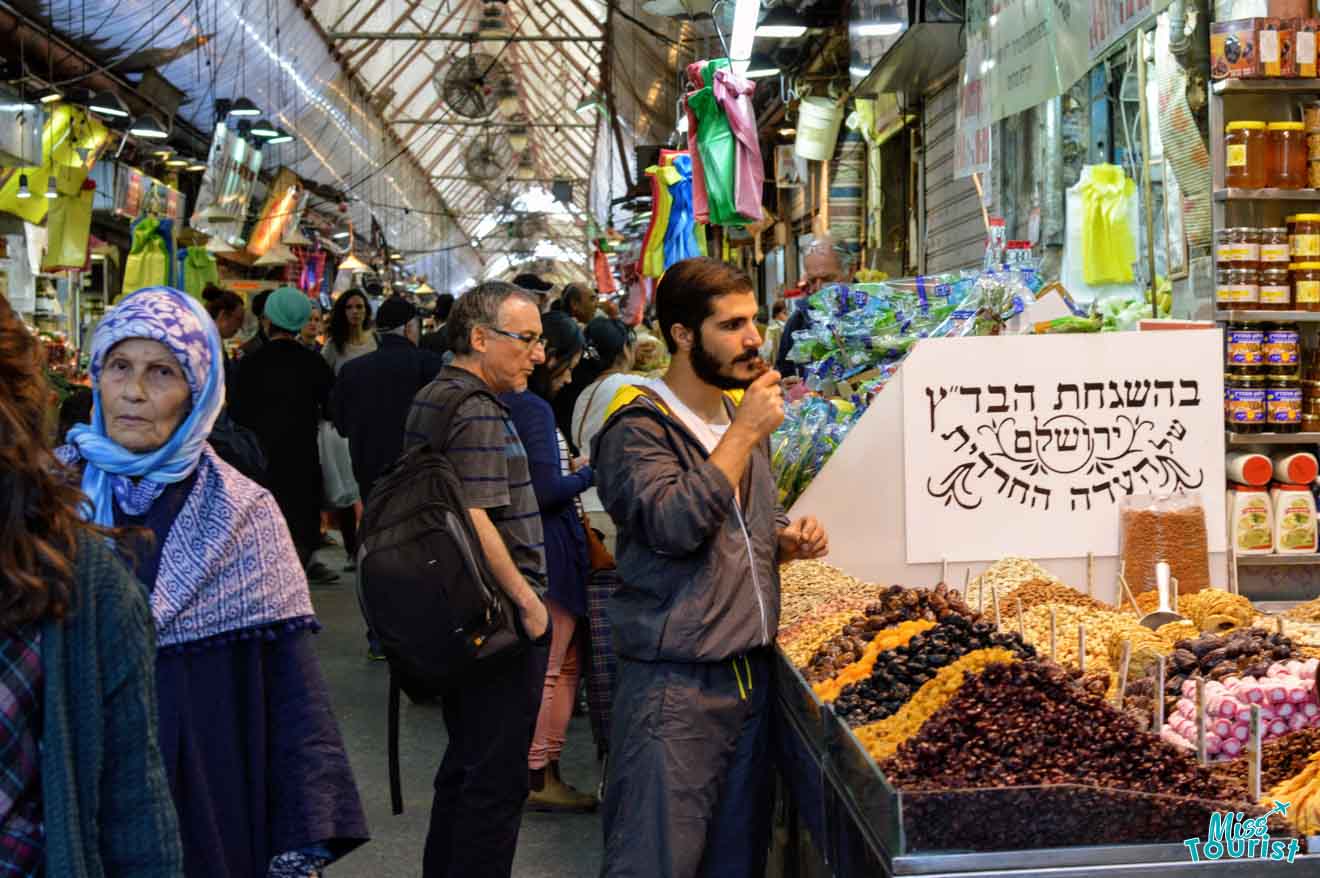 the author of this post visiting a bustling marketplace in Jerusalem with diverse shoppers, stalls with dried fruits and nuts, and a sign in Hebrew script.