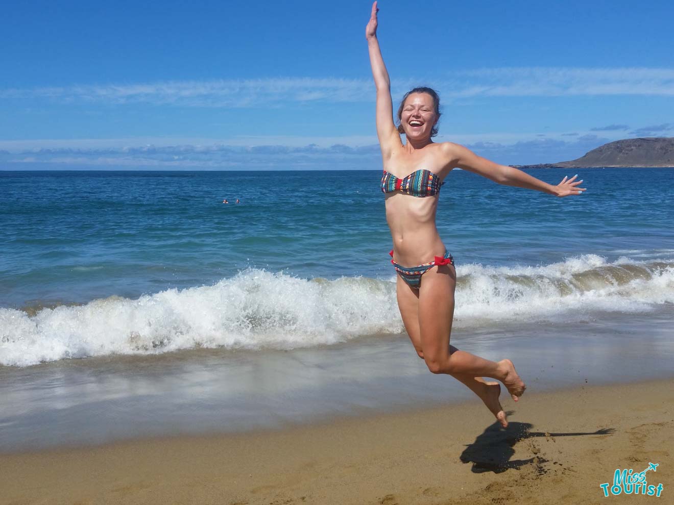 Author pf the post in a colorful bikini jumps in the air on a sandy beach with waves crashing behind them and a clear blue sky above.