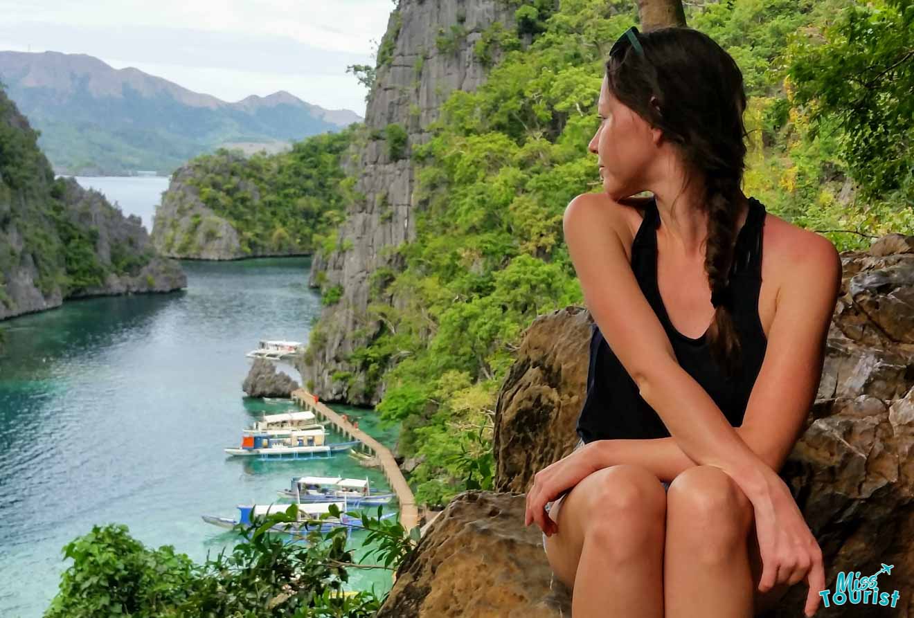 author of the post sits on a rock, looking over her shoulder at a scenic view of a narrow bay surrounded by steep, lush green cliffs with several boats anchored along the shore.