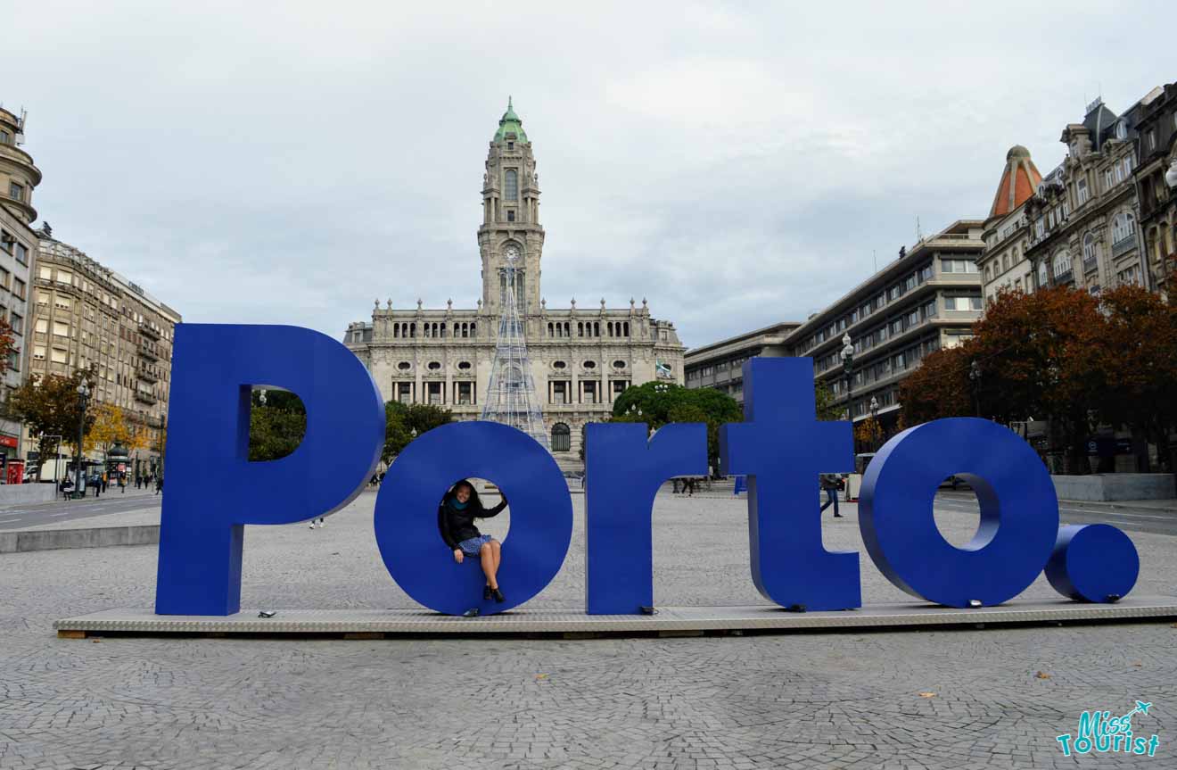 The iconic blue 'Porto.' sign in front of the ornate City Hall in Avenida dos Aliados, Porto, with the writer of the article sitting in the letter 'O', under a cloudy sky