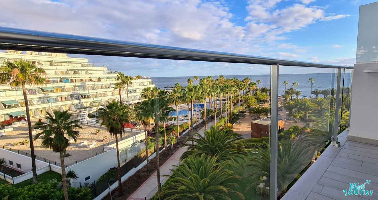 Balcony view from a hotel room showcasing a row of palm trees, swimming pools, and the ocean horizon at sunset