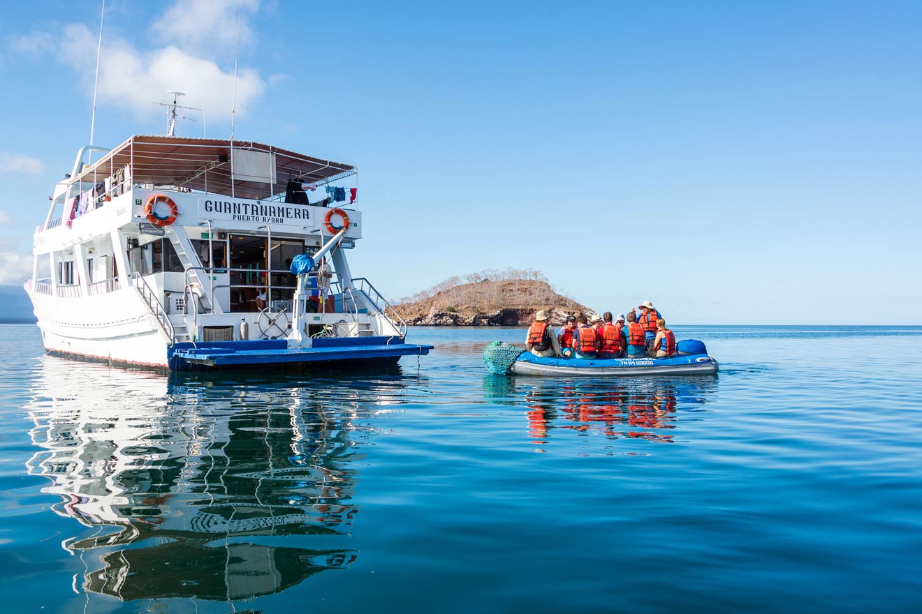 A group of people in life vests sit on a small inflatable boat near a larger boat named “Guantanamera” on calm water with an island in the background.