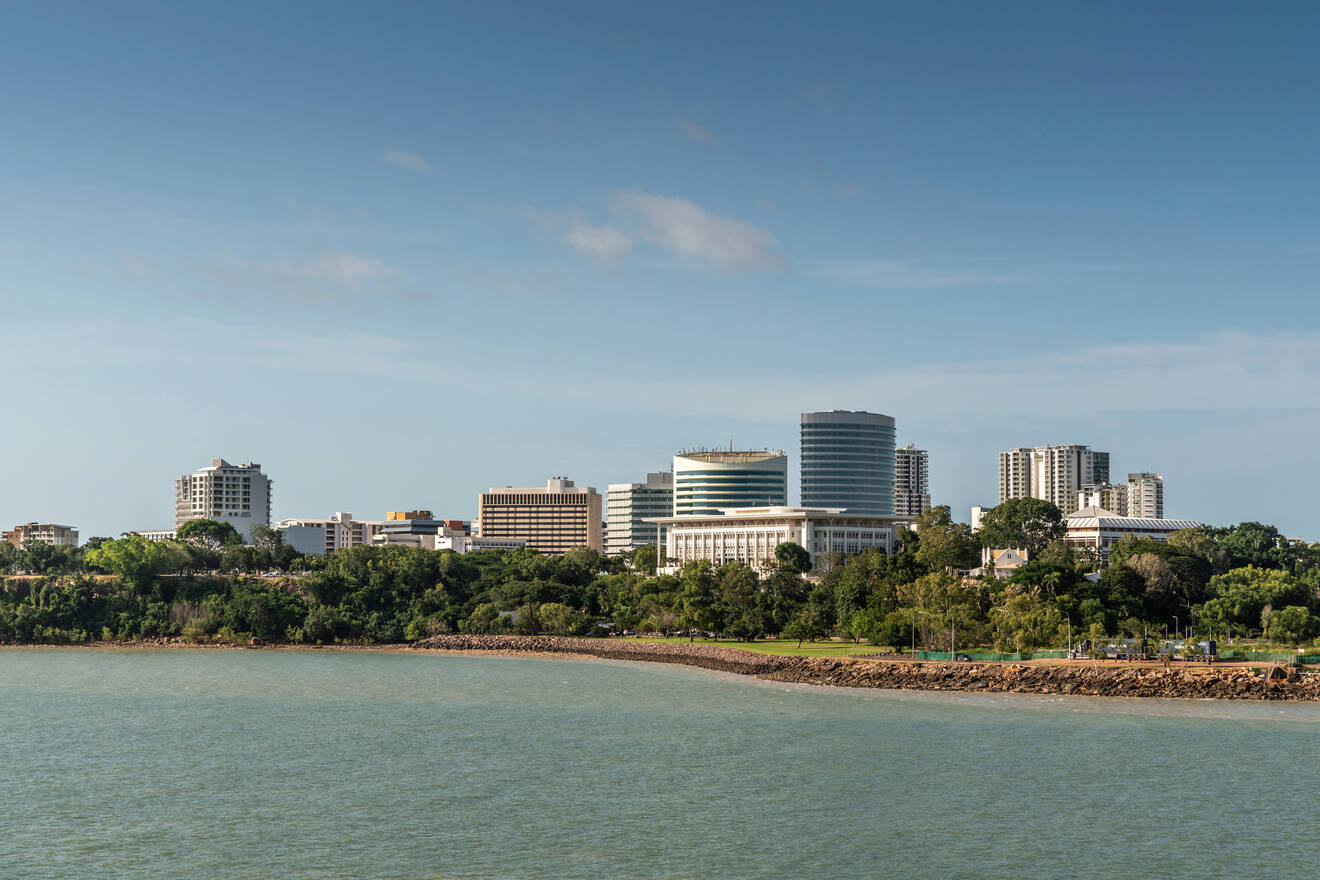 Skyline of a city with multiple mid-rise buildings near a body of water, with trees and greenery in the foreground.