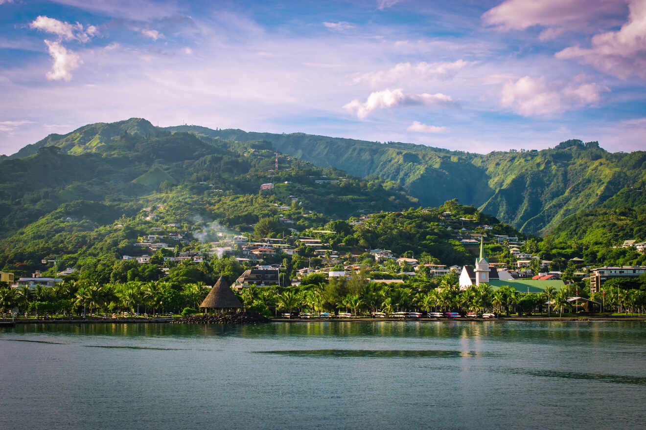 A scenic view of a lush green mountainside town in Tahiti with a church and a traditional thatched-roof building along the waterfront.