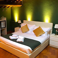A neatly made bed with white linens, green bed runner, and gold pillows in a room with green walls and wooden flooring, illuminated by warm lighting.
