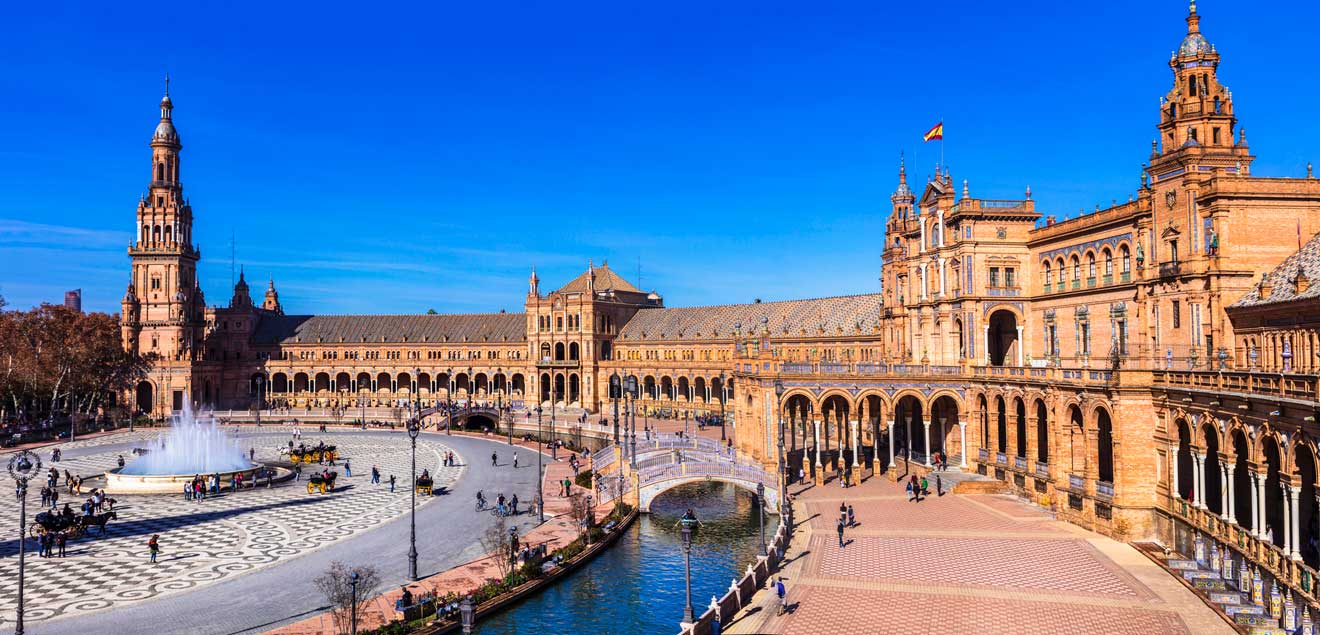 Panoramic view of Plaza de España in Seville, showcasing the magnificent Spanish Renaissance architecture, with a large fountain centering the semicircular plaza, and horse-drawn carriages by the canal under a clear blue sky