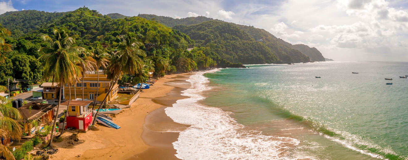 Aerial view of a scenic tropical beach with calm turquoise waters, lush green hills, and palm trees. Some colorful buildings and boats are visible along the shoreline.