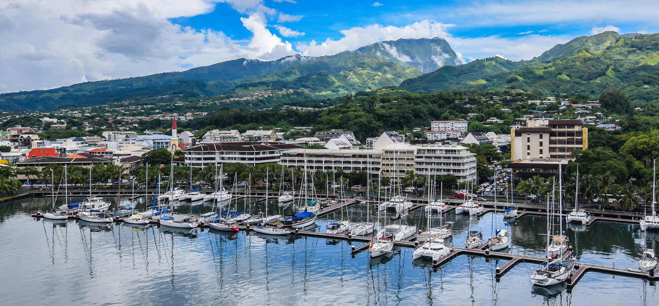 A marina with numerous sailboats docked, set against a backdrop of green mountains and a vibrant town in Tahiti