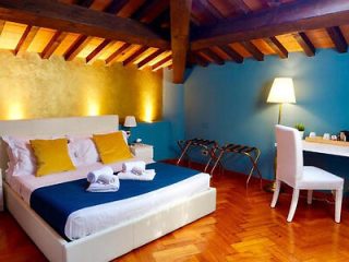 A neatly arranged bedroom with a large bed, blue blanket, and yellow pillows. The room features a wooden beamed ceiling, a desk with a chair, a lamp, and modern decor including a small nightstand.