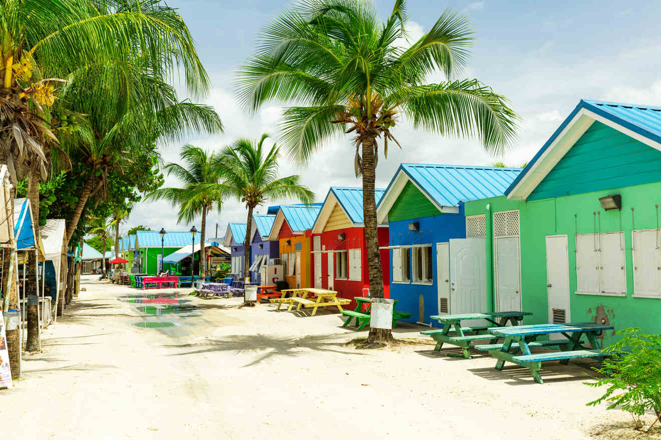 A row of colorful beach huts with palm trees lining a sandy walkway under a partly cloudy sky.