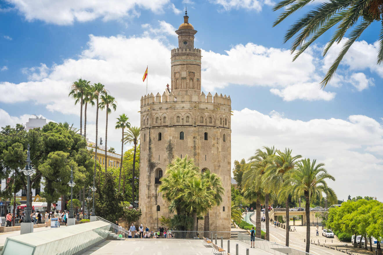 Iconic Torre del Oro, a dodecagonal military watchtower, under a bright blue sky adorned with clouds in Seville, Spain, flanked by lush palm trees and a busy promenade alongside the Guadalquivir River