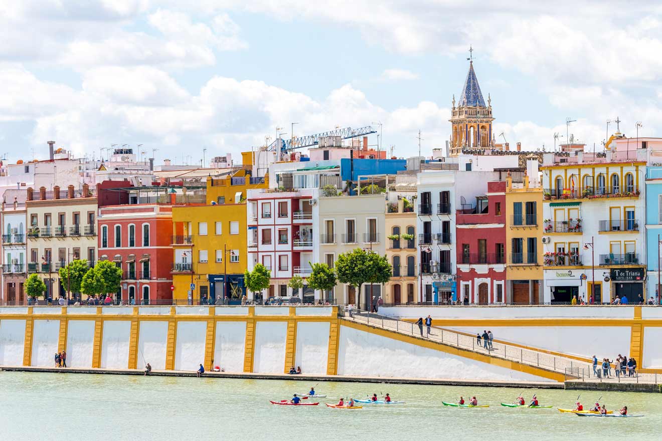 Kayakers enjoying a sunny day on the river against the backdrop of Seville's vibrant Triana neighborhood, with its colorful facades and the iconic tower of the Church of Santa Ana rising in the distance