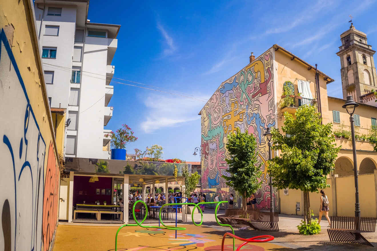 A vibrant urban courtyard features colorful street art on building walls, modern sculptures, and outdoor seating areas with people enjoying the sunny day.
