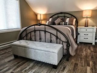 A bedroom with an arched metal bed, gray bedding, matching nightstands, and lamps on either side. A cushioned bench sits at the foot of the bed on a wooden floor.