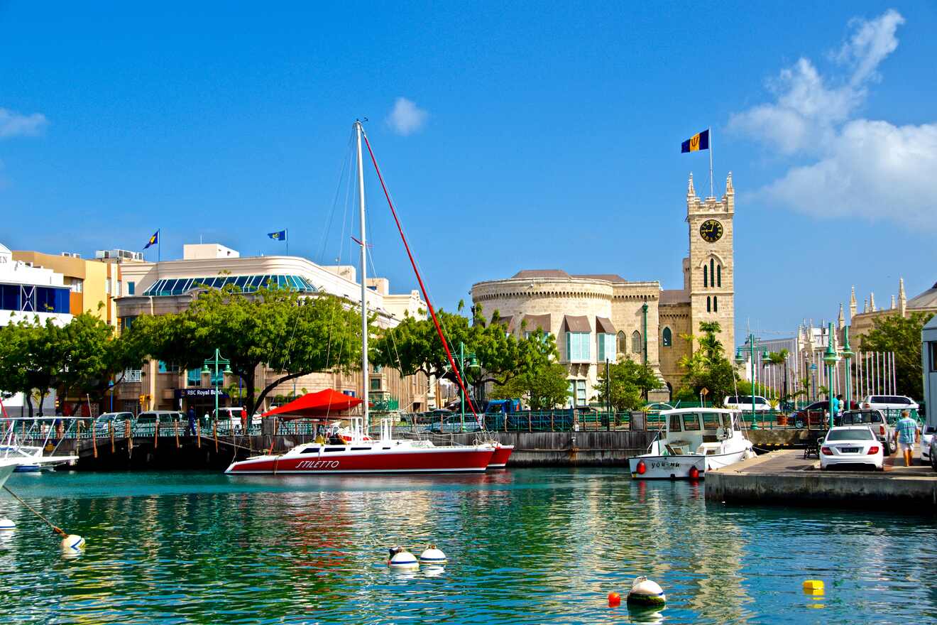 A picturesque harbor with yachts docked in clear water, a historic clock tower, and buildings in the background under a bright blue sky.