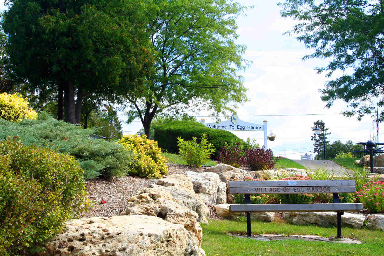 A landscaped park with a wooden bench labeled "Village of Egg Harbor," surrounded by rocks, bushes, and trees. A sign in the background reads "Welcome to Egg Harbor.