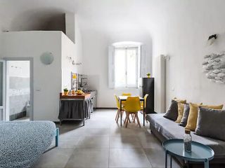 Modern studio apartment with a kitchenette, dining area, sofa, and bed. The space features minimalist decor with white walls, light gray floors, and a mix of yellow and neutral accents.