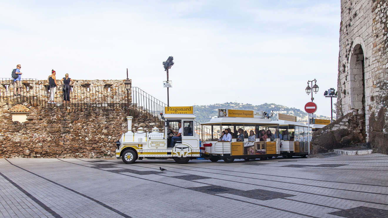 A white tourist train with yellow accents and passengers is seen near an ancient stone structure. People stand on the structure, and hills are visible in the background.