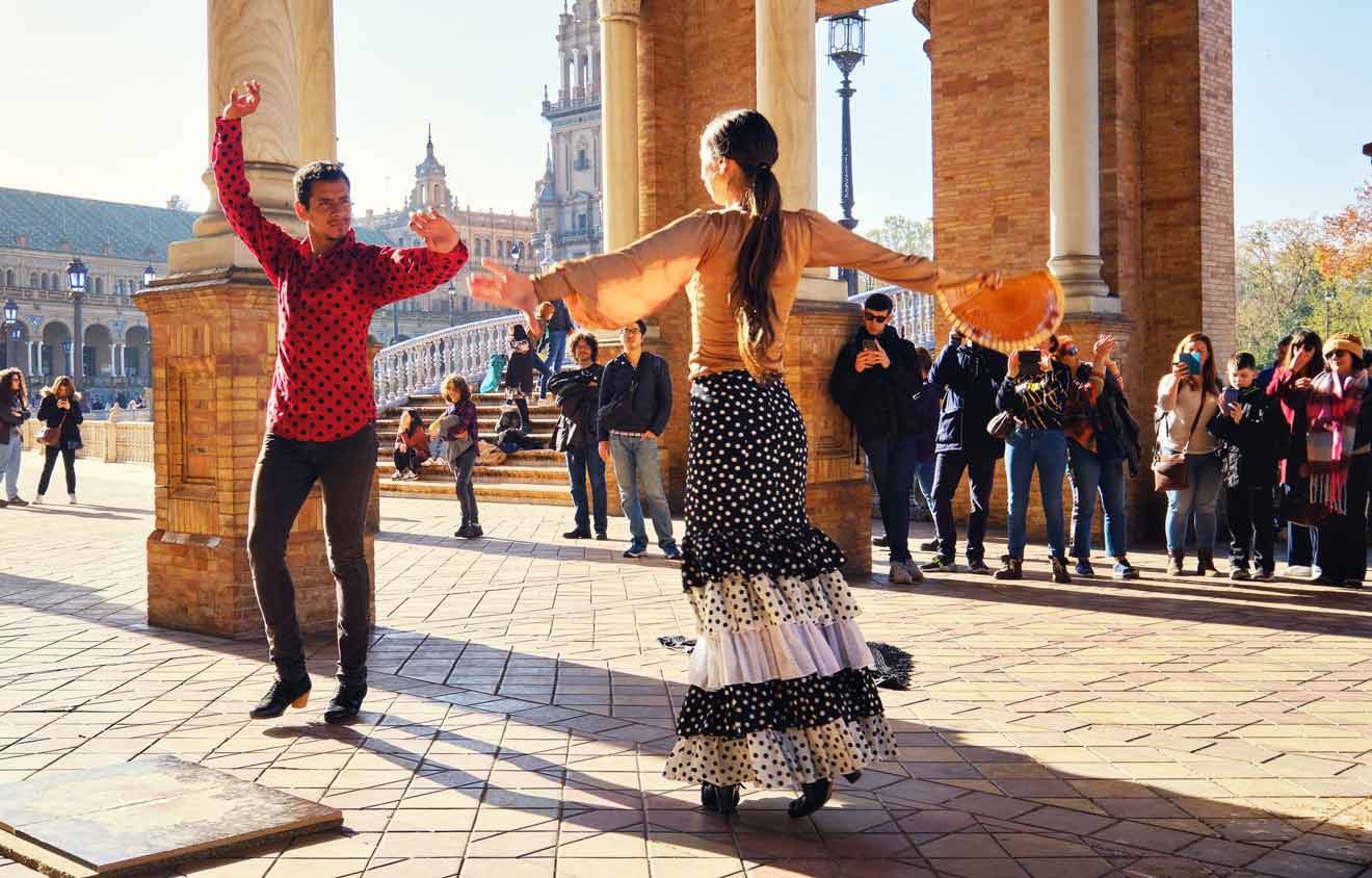 A flamenco dancer in traditional polka dot attire and her partner perform an impassioned dance at Seville's Plaza de España, surrounded by an audience captivated by the vibrant display of Andalusian culture