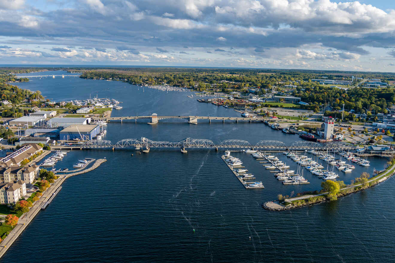 Aerial view of a seaside town featuring a bridge, marinas with docked boats, and numerous buildings along the waterfront under a partly cloudy sky.