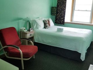 A small, tidy bedroom with a single bed, red armchair, phone, and table lamp. The walls are painted light green, and there's a window with patterned curtains. A note is placed on the bed.
