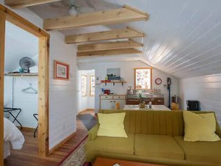 A cozy room with wooden beams, a green sofa with yellow pillows, white walls, a small kitchen area, and a doorway to a bedroom with a visible bed and closet. Natural light enters through two windows.