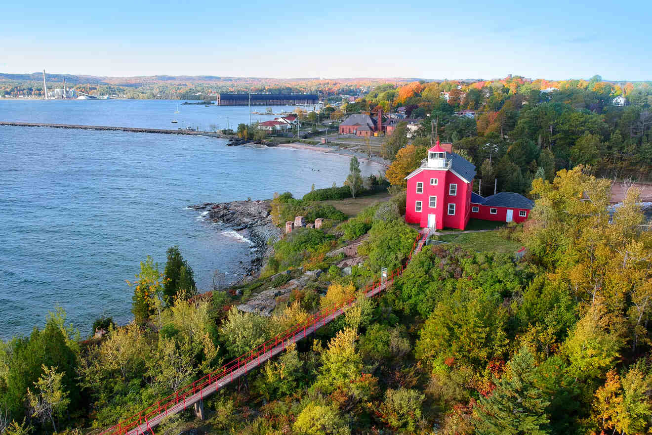 A red lighthouse on a grassy hill by the shore, surrounded by trees with autumn foliage, overlooking a calm body of water.