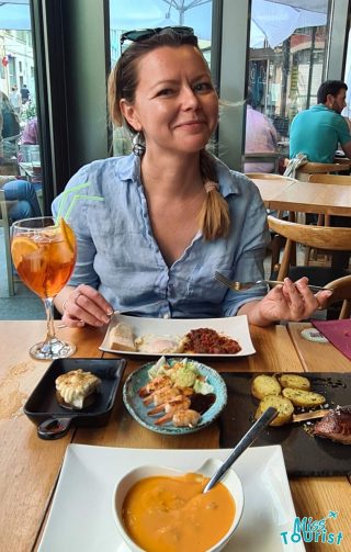 Dining experience in a casual restaurant, featuring a smiling woman with a spread of Spanish cuisine, including a bowl of creamy soup, assorted tapas, and a refreshing Aperol Spritz, embodying the leisurely and flavorful essence of Spanish dining