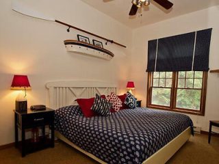 A bedroom with a nautical theme, featuring a king-sized bed with patterned bedding, red lamps on bedside tables, and a canoe shelf above the headboard. A window with dark blue curtains is on the right.