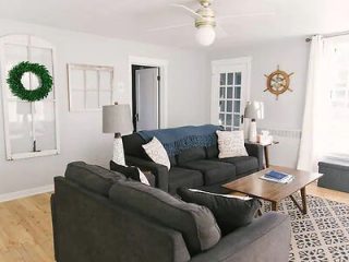 A bright living room featuring a dark grey couch, wooden coffee table, lamp, nautical decor, and large white-framed windows. A green wreath hangs on one wall, with light wood flooring throughout.