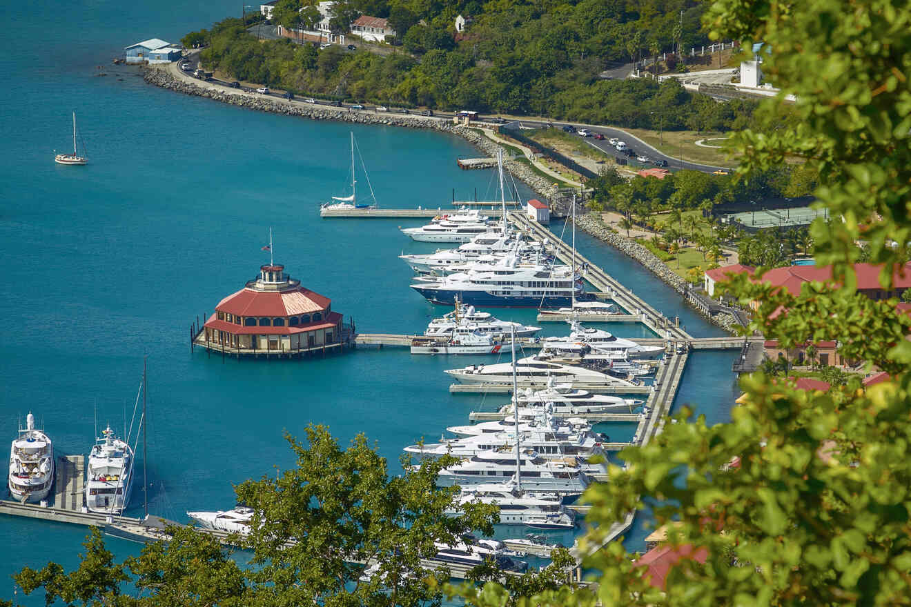 Aerial view of a marina with multiple docked yachts, a circular building in the center, and surrounding green foliage by clear blue water.