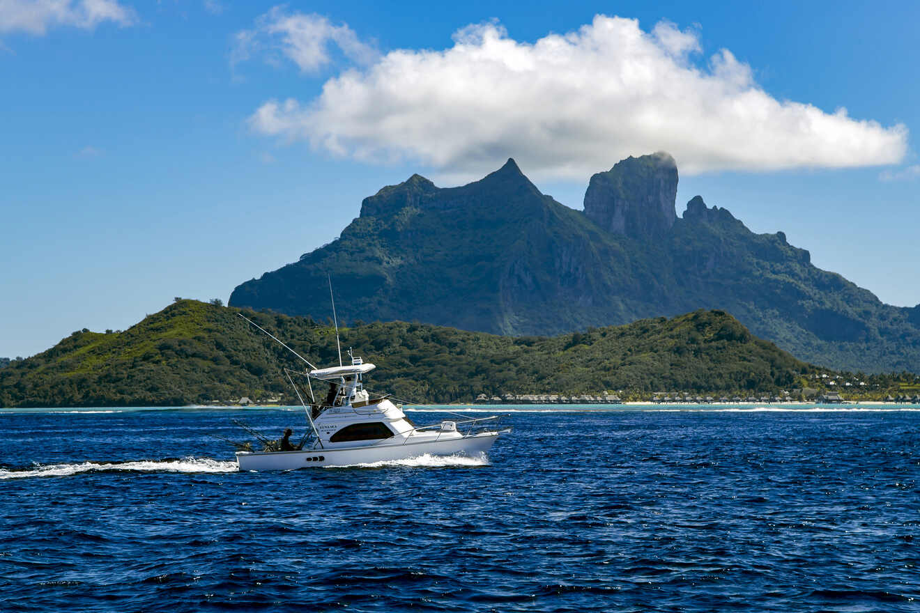 A fishing boat cruising on the blue ocean with a rugged green mountain and cloud-covered peaks in the distance.