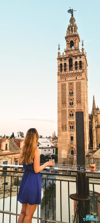 A woman enjoys a serene moment on a balcony with a drink in hand, overlooking the magnificent Giralda Tower in Seville, as the golden hues of sunset cast a warm glow over the historic architecture