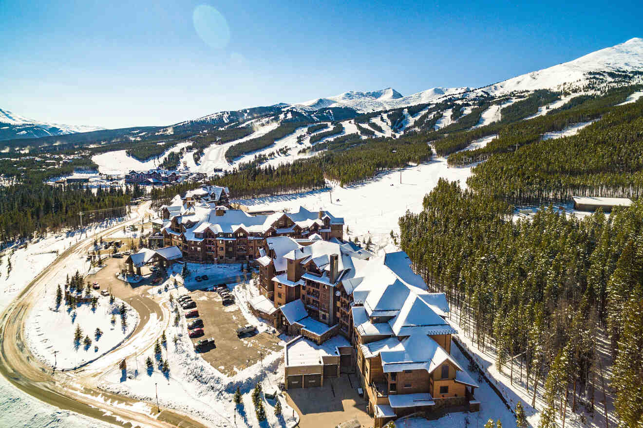 Aerial view of a snowy mountain resort with buildings surrounded by pine trees, winding road, parking area, and ski slopes in the background. Clear blue sky overhead.